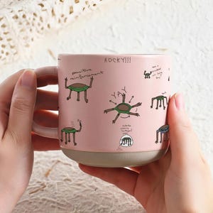 May include: A pink ceramic mug with a light beige base and handle. The mug is decorated with cartoon drawings of creatures and handwritten text, including "ROCKY!!!" and "positive reinforcement."