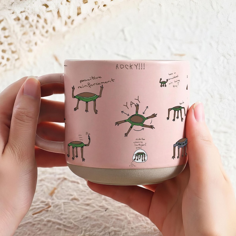 May include: A pink ceramic mug with a light beige base and handle. The mug is decorated with cartoon drawings of creatures and handwritten text, including "ROCKY!!!" and "positive reinforcement."