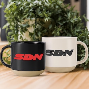 May include: Two ceramic mugs, one black and one off-white, with a beige base. The black mug features the red letters "SDN" with a starburst design. The off-white mug has the same logo in black. Both mugs have a curved handle.