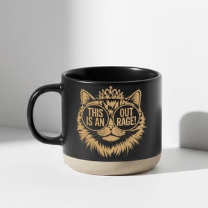 May include: Black ceramic mug with a tan base and a black handle. The mug features a gold illustration of a cat wearing sunglasses and a crown, with the text "THIS IS AN OUT RAGE!" in gold.