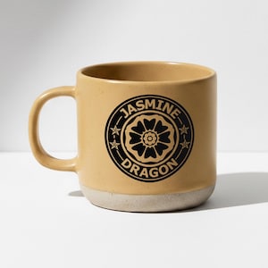 Könnte beinhalten: Eine beige Keramiktasse mit rundem Henkel und weißem Boden. Die Tasse hat ein schwarzes kreisförmiges Design mit den Worten "Jasmine Dragon" und einem floralen Emblem.