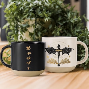 Peut inclure: Deux mugs en céramique. L'un est noir avec des silhouettes de dragons dorés. L'autre est blanc avec un motif de dragon noir, des accents dorés et une base beige. Les deux mugs ont une anse incurvée et une base beige.