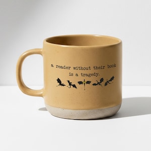 Op de afbeelding: Tan keramische mok met een witte basis. De mok heeft de tekst "a reader without their book is a tragedy" in zwarte tekst, en zwarte draken silhouetten. De mok heeft een groot handvat.