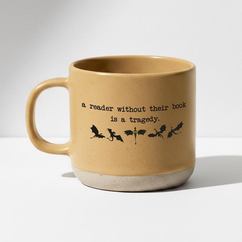 May include: Tan ceramic mug with a white base. The mug features the quote "a reader without their book is a tragedy" in black text, along with black dragon silhouettes. The mug has a large handle.