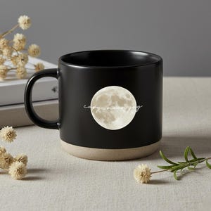 Könnte beinhalten: Schwarze Keramik-Tasse mit einem Vollmond-Motiv und dem Schriftzug "copy, moon, joy". Der Boden der Tasse ist hellbeige. Die Tasse hat einen Henkel und steht auf einer neutralen Oberfläche. Geeignet für Heißgetränke.