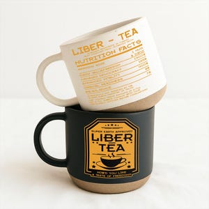 Könnte beinhalten: Zwei Keramikbecher übereinander gestapelt. Der obere Becher ist weiß mit einem beigefarbenen Boden und dem Text "Liber Tea - Nutrition Facts". Der untere Becher ist schwarz mit beigefarbenem Boden und einem goldenen Etikett "Liber Tea".