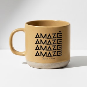 Könnte beinhalten: Eine beige Keramik-Kaffeetasse mit einem abgerundeten Henkel. Die Tasse zeigt das Wort "AMAZE" viermal in Schwarz gestapelt, mit einer kleinen Illustration über dem Wort. Der Boden der Tasse ist heller, und die Worte "Ryland & Rocky" sind darunter gedruckt.