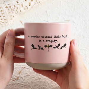 May include: A pink ceramic mug with a gray base and handle. The mug features the quote "a reader without their book is a tragedy" and dragon silhouettes. The mug is held by two hands.