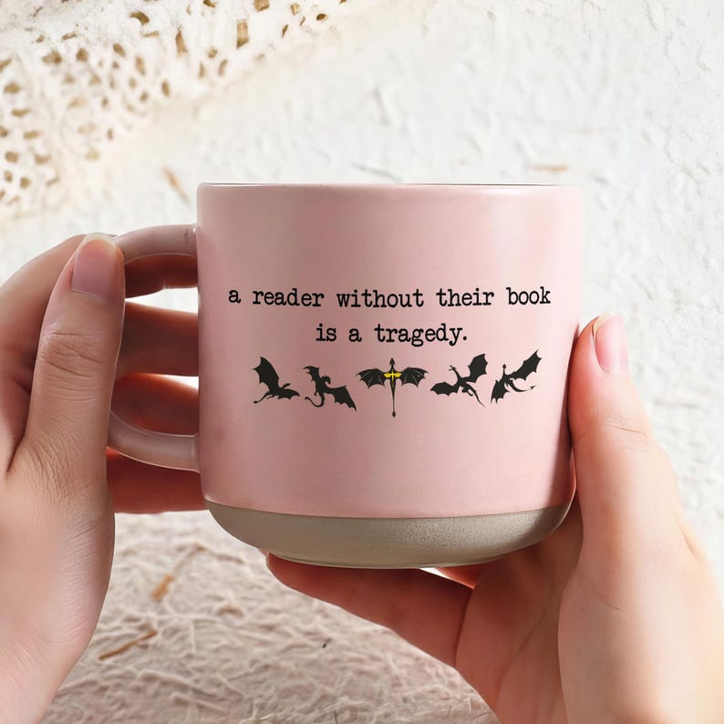 May include: A pink ceramic mug with a gray base and handle. The mug features the quote "a reader without their book is a tragedy" and dragon silhouettes. The mug is held by two hands.