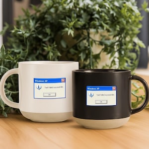 Pode incluir: Duas canecas de café de cerâmica, uma branca e outra preta, com base bege. Cada caneca apresenta uma mensagem de erro do Windows XP: "Task failed successfully." As canecas têm um design clássico com alça e estão sobre um fundo verde desfocado.
