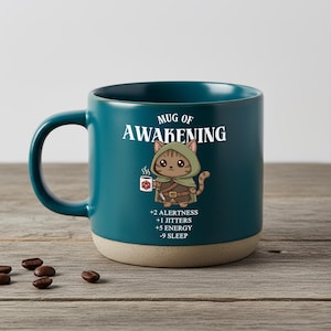 Op de afbeelding: Teal keramische mok met een beige basis, met de tekst "Mug of Awakening" en een cartoon kat karakter met een koffiekopje. De mok heeft een gebogen handvat.