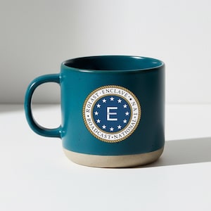 May include: A white ceramic mug with a black handle and the text "But First, Coffee" in a bold, black font. The mug is set against a blurred background, highlighting its simple design.