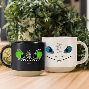 May include: Two ceramic mugs featuring dragon face designs. One mug is black with green eyes, the other is white with blue eyes. Both mugs have a beige base and a handle. The mugs are on a wooden surface.