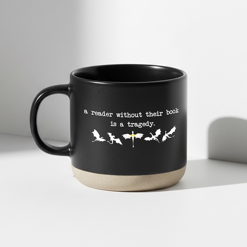 May include: Black ceramic mug with a beige base and a black handle. The mug features the text "a reader without their book is a tragedy." and a row of white dragon silhouettes.