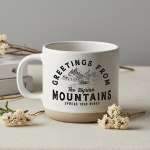 May include: White ceramic mug with a beige base, featuring the text "GREETINGS FROM The Illyrian MOUNTAINS" in black. The design includes a mountain illustration. The mug has a curved handle and is surrounded by white flowers.