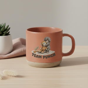 May include: A coral-colored ceramic mug with a light beige base. The mug features a graphic of two cartoon monkeys hugging, with the text "TEAM PUNCH" below. The mug is on a light wooden surface.