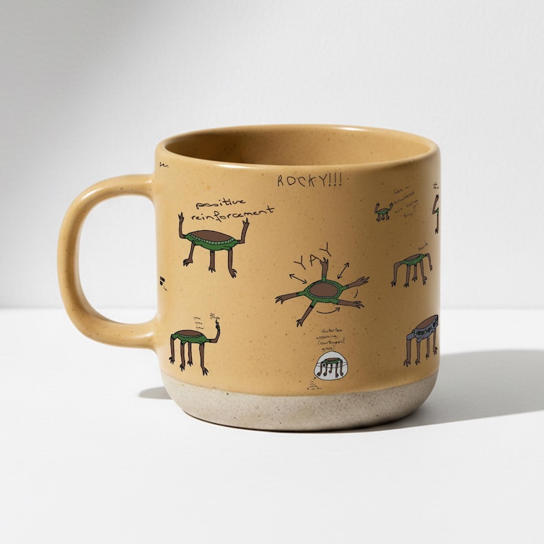 May include: Tan ceramic mug with a speckled finish, featuring whimsical green and brown creature illustrations and the phrases "positive reinforcement" and "ROCKY!!!!". The mug has a beige base and a curved handle.