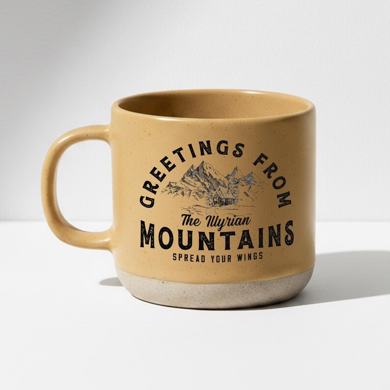 May include: A tan ceramic mug with a white base and a curved handle. The mug features the text "GREETINGS FROM The Illyrian MOUNTAINS" with a mountain graphic. The phrase "SPREAD YOUR WINGS" is printed below the mountain graphic.