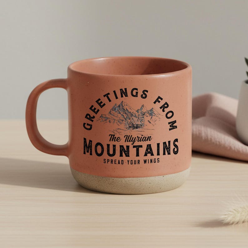 May include: A white ceramic mug with a black handle and the text "But First, Coffee" in a bold, black font. The mug is set against a blurred background. The mug appears to be approximately 10 cm tall.
