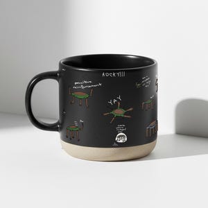 May include: A white ceramic mug with a black handle and the text "But First, Coffee" in a black, handwritten-style font. The mug is on a white surface.