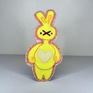May include: A yellow felt bunny-shaped decoration with a white heart on its chest. The bunny has a stitched X for a mouth and a pink outline with beaded trim. The ears are upright and the arms are rounded.