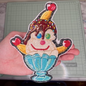 May include: Embroidered patch featuring a whimsical ice cream sundae design. The patch showcases a scoop of ice cream with chocolate sauce, sprinkles, and a cherry on top. The sundae is in a blue sundae dish, with a smiling face.