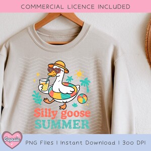 K&ouml;nnte beinhalten: Ein hellbeiger Pullover mit einem Grafikdesign eines Cartoon-G&auml;nses, der eine Sonnenbrille und einen Hut tr&auml;gt, ein Glas Saft h&auml;lt und auf einem bunten Schwimmring schwimmt. Der Text "Silly goose SUMMER" ist unter dem Gans gedruckt.