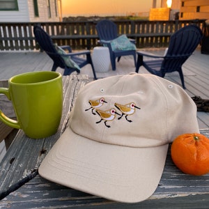 Puede incluir: Una gorra de béisbol beige con un diseño bordado de cuatro pájaros amarillos con picos rojos caminando en línea. La gorra está sentada sobre una superficie de madera junto a una taza verde y una naranja.