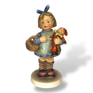 May include: A porcelain figurine of a young girl holding a doll and a basket. The girl wears a blue bow, a green and blue dress, and brown boots. The doll has a red coat and yellow collar. The figurine is on a round, off-white base.
