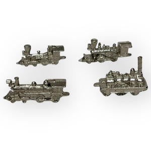 May include: Four small, silver-colored train miniatures are arranged on a white background. Each miniature train has detailed features, including a smokestack, wheels, and a cab. The trains vary slightly in design, showcasing different locomotive styles.