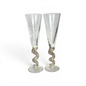 May include: Two clear champagne flutes with textured glass and a unique, swirled stem design. The stems have a textured, metallic appearance, and the flutes sit on a round base. The glasses are set against a white background.