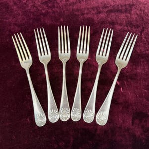 May include: A set of six silver-toned metal forks arranged in a fan shape. Each fork features four long tines and a handle decorated with a raised, embossed shell-like motif at the base. The metal has a polished, reflective finish throughout.