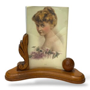 May include: A vintage wooden picture frame with a portrait of a woman. The frame has a curved base with a decorative carved element on the left and a small wooden sphere on the right. The portrait features a woman with light skin, and a floral arrangement.