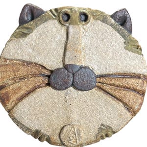 May include: A circular ceramic wall plaque featuring a stylized cat face. The piece is crafted from textured, earthy-toned clay with dark brown accents on the ears, nose, and whiskers. A small circular stamp with an embossed symbol is at the bottom.
