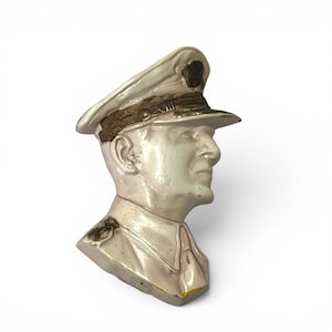 May include: A vintage, off-white bust of a man in a military cap, viewed in profile. The bust has a detailed face and uniform, with a weathered appearance. The cap features a dark emblem and a gold-colored band.