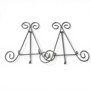 May include: A decorative metal display stand with a gray finish. The stand features two easel-style supports connected by a central, looped design. Each support has a scrolled top and a small shelf.