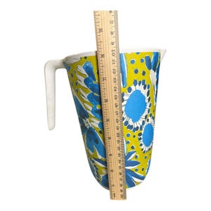Op de afbeelding: Een vintage kan met een witte handgreep en een levendig bloemenpatroon in blauw en wit op een gele achtergrond. De kan is ongeveer 25 cm hoog, gemeten met een liniaal.