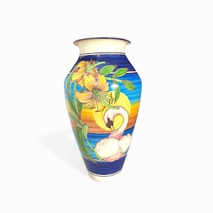 May include: A decorative ceramic vase with a hand-painted design. The vase features a swan, yellow lilies, and a sunset scene. The vase has a blue rim and base, with a white top.