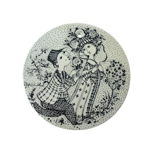 May include: A round white ceramic ornament featuring a black ink illustration of a person kneeling to offer flowers to another person. The design includes floral motifs, a small bird, and the signature Bjørn Wiinblad printed on the left side.