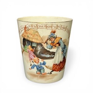 May include: A cream-colored ceramic mug featuring a vintage illustration of a woman with a broom and children near a shoe-shaped house. The text "There was an Old Woman who lived in a shoe" is printed in a banner across the top of the decorative vessel.