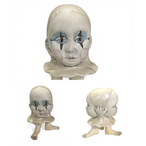 May include: A ceramic sculpture of a doll-like head with painted facial features, including blue eyeshadow and pink blush. The head is attached to a ruffled collar and small legs. The sculpture has a distressed, aged appearance.