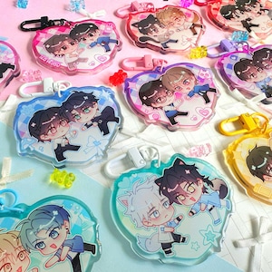 May include: A collection of heart-shaped acrylic keychains featuring colourful anime-style character illustrations. Each keychain includes a plastic clasp and displays two figures in various poses, accented with small bows and scattered star motifs.