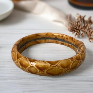 May include: A round, brown bangle bracelet with a reptile-like pattern. The bracelet has a dark interior and a glossy finish. The background includes a white surface and blurred objects.