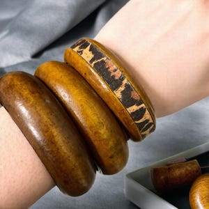 May include: Three wooden bangles in varying shades of brown are stacked on a wrist. The top bangle features a decorative band with a brown and tan animal print. The other two bangles are solid brown.