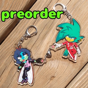 Peut inclure: Deux porte-clés en acrylique transparent avec des personnages de dessins animés. L'un représente un personnage aux cheveux bleus dans une tenue rose et noire, l'autre un personnage aux cheveux verts dans une combinaison de course rouge. Le mot "preorder" est en vert au-dessus.