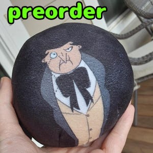 Puede incluir: Un juguete de peluche negro y blanco de un personaje con una pajarita negra y una camisa blanca. El personaje tiene una nariz grande y una expresión seria. El juguete es redondo y tiene una superficie texturizada. El texto "preorder" está escrito en letras verdes sobre el juguete.