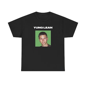 Puede incluir: Camiseta negra con el texto "YUNG LEAN" sobre un retrato de un hombre con fondo verde. La camiseta es de cuello redondo.