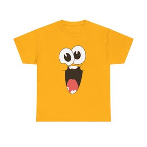 May include: Mustard yellow t-shirt featuring a cartoon face with large, wide eyes, a wide open mouth, and a pink tongue. The graphic is centered on the front of the shirt. The shirt is made of a soft material.