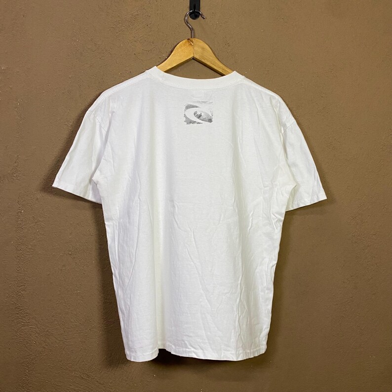 May include: White short-sleeve t-shirt hanging on a wooden hanger. The back of the shirt features a small, faded graphic design near the neckline. The shirt appears to be made of a soft, lightweight fabric, suitable for casual wear.