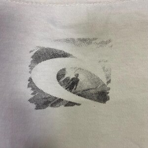 May include: Close-up of a white t-shirt featuring a black and white graphic design. The design is a stylized wave with a surfer silhouette inside a square frame. The shirt has a crew neck.